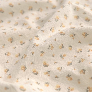 May include: Close-up of a cream-colored fabric with a delicate floral pattern. Small yellow flowers with green leaves are scattered across the surface. The fabric has a slightly wrinkled texture, suggesting it's soft and lightweight.