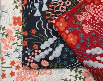May include: Three fabric swatches featuring embroidered floral designs. The top swatch is red with orange flowers, the middle swatch is navy blue with white, orange, and green flowers, and the bottom swatch is white with pink and green flowers.