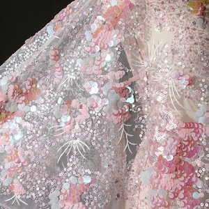 Può includere: Primo piano di un tessuto rosa e bianco con paillettes e un delicato motivo floreale. Le paillettes sono iridescenti e creano un effetto scintillante.