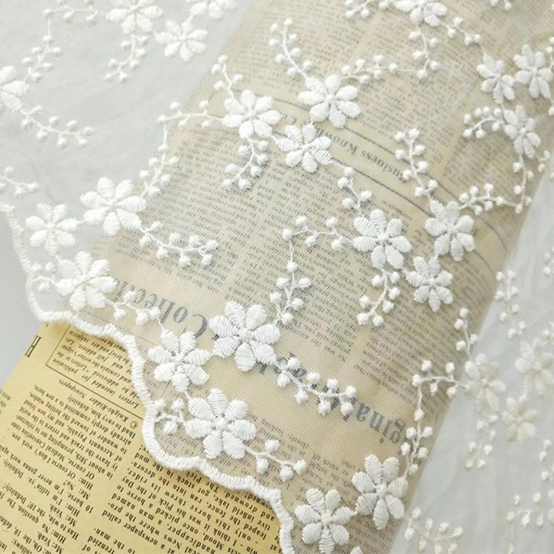 May include: White floral lace trim with a delicate pattern of small flowers and leaves. The lace is on a white background and has a scalloped edge.