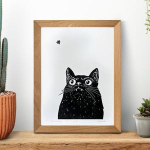 May include: Black and white linocut print of a cat looking up at a fly. The print is framed in a natural wood frame. The text "The Watching of a Time" is printed below the image.