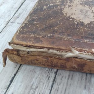 May include: Close-up of an antique book with a worn, brown leather cover. The edges of the pages are visible, and the binding shows signs of age and wear. The book is placed on a light-coloured wooden surface.