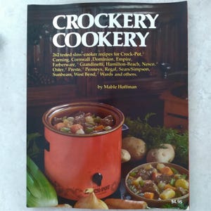 以下が含まれることがあります： メイブル・ホフマン著の料理本『Crockery Cookery』。スロークッカーのレシピ262品を掲載。表紙には、シチューが入った鮮やかなオレンジ色のスロークッカーと、ジャガイモ、ニンジン、リーキなどの新鮮な野菜が暗い背景に写っている。