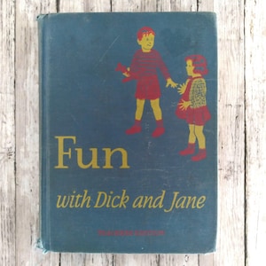 Pode incluir: Livro azul vintage intitulado "Fun with Dick and Jane". A capa apresenta ilustrações de duas crianças vestidas com roupas vermelhas e amarelas. O título é amarelo, com os nomes dos autores abaixo.