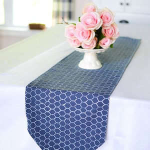 May include: A blue and white table runner with a chicken wire pattern. The runner is on a white tablecloth and has a bouquet of pink roses in a white pedestal vase.