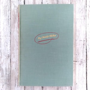 May include: A vintage book titled "My Friend Flicka" with a light teal cover. The title is in gold script inside a red oval. The book is set against a white and grey wood-grain background.