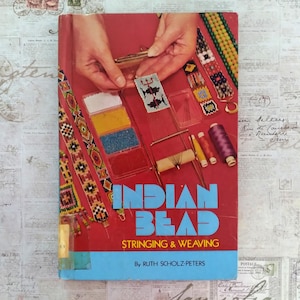 Może przedstawiać: Książka w stylu vintage zatytułowana "Indian Bead Stringing & Weaving" autorstwa Ruth Scholz-Peters. Okładka jest czerwona z niebieskim tekstem i zawiera zdjęcia rękodzieła z koralików, szpulek nici i rąk pracujących z koralikami.