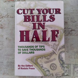 May include: A book titled Cut Your Bills In Half by the Editors of Rodale Press. The white cover features large maroon text and a graphic of green 1000 dollar bills. The subtitle reads Thousands of Tips to Save Thousands of Dollars in green.