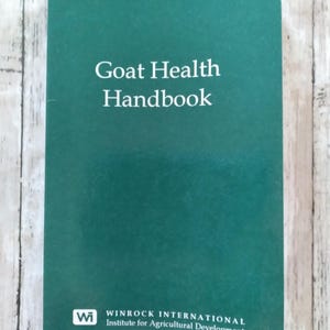 May include: A green handbook titled "Goat Health Handbook" with white text. The cover also includes the Winrock International logo and Institute for Agricultural Development text. The handbook is on a light wood grain surface.