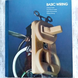 Peut inclure: Un livre intitulé "BASIC WIRING" avec une couverture bleue. La couverture présente un gros plan d'une prise et d'un interrupteur électriques beiges, avec un câblage et un tournevis visibles. Le texte sur la couverture comprend des sujets tels que l'ajout de prises et le câblage extérieur.