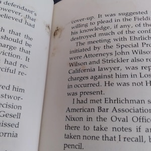 May include: An open book with off-white pages featuring black printed text. The left page shows partial sentences, while the right page contains a paragraph discussing a meeting with Ehrlichman and the American Bar Association. There are small brown stains.