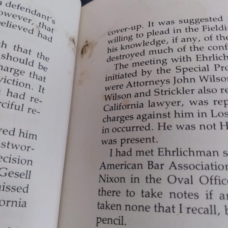May include: An open book with off-white pages featuring black printed text. The left page shows partial sentences, while the right page contains a paragraph discussing a meeting with Ehrlichman and the American Bar Association. There are small brown stains.