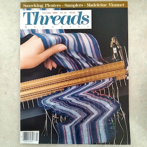 May include: Threads Magazine issue No. 21 from Feb/Mar 1989 featuring a gold-toned metal smocking pleater tool. The cover displays a blue, white, and magenta striped fabric being fed through the pleater, creating a distinct chevron pattern.