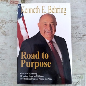 May include: A book titled Road to Purpose by Kenneth E. Behring. The cover features a photograph of a smiling man in a dark suit and red tie, with an American flag in the background. The text reads: Road to Purpose, One Man's Journey Bringing Hope to Millions and Finding Purpose Along the Way.