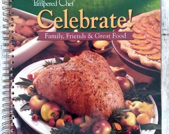 May include: A spiral-bound cookbook titled "Celebrate!" by The Pampered Chef. The cover features a roasted turkey surrounded by apples and cranberries, with a fruit tart in the background. The text reads "Family, Friends & Great Food."