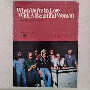 Puede incluir: Una partitura vintage titulada When You're In Love With A Beautiful Woman de Even Stevens, grabada por Dr. Hook. La mitad superior es de color rojo oscuro con texto blanco, y la inferior muestra una foto de una banda de seis hombres.