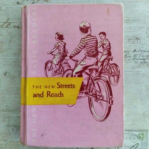 Könnte beinhalten: Vintage Kinderbuch mit dem Titel "The New Streets and Roads". Das Cover ist rosa mit Illustrationen von Kindern auf Fahrrädern in Kastanienbraun. Der Titel befindet sich auf einem gelben Band. "Basic Readers" ist vertikal auf der linken Seite gedruckt.