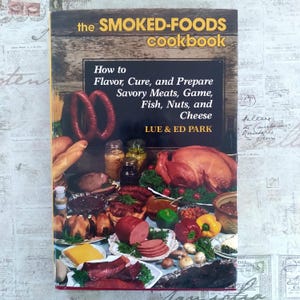 Peut inclure: Un livre de cuisine intitulé "the SMOKED-FOODS cookbook" avec une couverture en bois. La couverture présente une exposition de viandes fumées, de poissons, de noix et de fromage. Le titre et le sous-titre du livre sont en gras, avec le nom de l'auteur en bas.