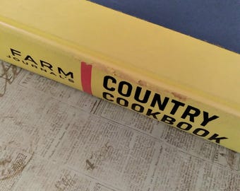May include: A hardcover book with a yellow spine featuring the black text Farm Journal's Country Cookbook. A thin vertical red stripe separates the words. The top edge of the spine shows signs of wear and minor paper loss along the yellow surface.