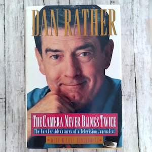 Pode incluir: Capa de livro com Dan Rather e o título "DAN RATHER" em letras douradas. O livro é intitulado "THE CAMERA NEVER BLINKS TWICE" e subtitulado "The Further Adventures of a Television Journalist".