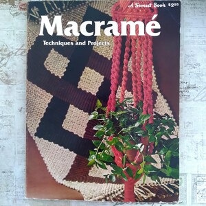 May include: A vintage book cover titled "Macramé: Techniques and Projects." The cover features a red macramé plant hanger holding a potted plant with green leaves. The background shows a macramé wall hanging with brown, black, and cream colors.