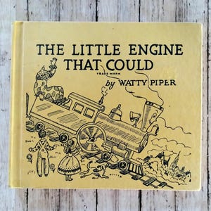 Könnte beinhalten: Ein Vintage-Kinderbuch mit dem Titel "The Little Engine That Could" von Watty Piper. Das Cover zeigt eine Schwarz-Weiß-Illustration einer lächelnden Lokomotive, eines Clowns, Kindern und eines Hundes. Das Buch hat einen hellgelben Einband.