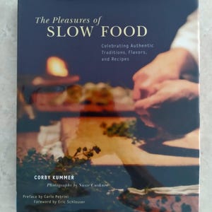 May include: A dark blue hardcover book titled "The Pleasures of Slow Food" with the subtitle "Celebrating Authentic Traditions, Flavors, and Recipes." The cover features a blurred image of food preparation. The author is Corby Kummer.