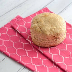 以下が含まれることがあります： ピンクと白の模様の布製のナプキンの上に置かれた、丸い薄い茶色のビスケット1つ