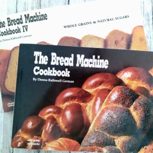 May include: Two cookbooks titled "The Bread Machine Cookbook" and "The Bread Machine Cookbook IV" are displayed. The covers feature images of various breads. The text on the covers includes the author's name, Donna Rathmell German, and the phrase "Whole Grains & Natural Sugars."