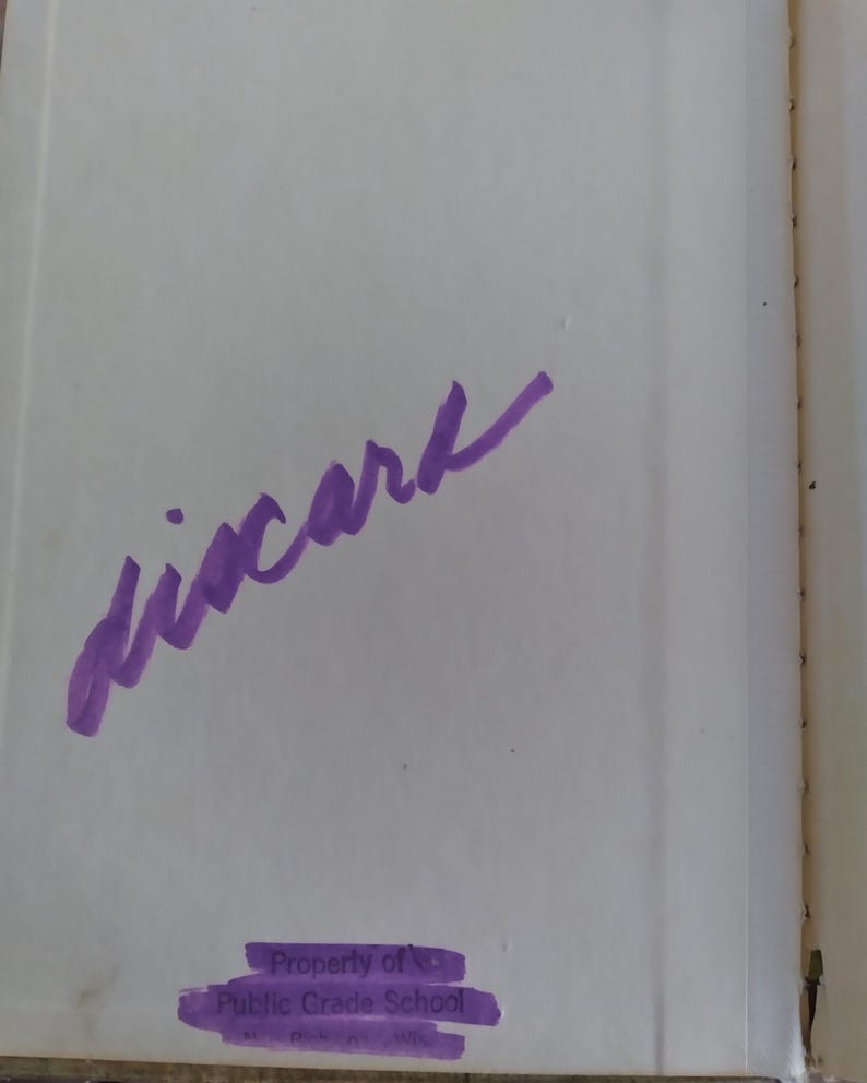 Pu&ograve; includere: Pagina di libro aperta con testo manoscritto viola che recita "xicark". Sotto il testo c'&egrave; un rettangolo viola con la scritta "Property of" e "Public Grade School".