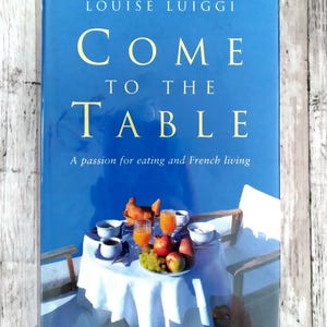 Könnte beinhalten: Ein Buch mit dem Titel "COME TO THE TABLE" von Louise Luiggi. Das Cover ist blau mit goldfarbenen Buchstaben und zeigt einen Tisch mit Essen und Getränken. Der Untertitel lautet "A passion for eating and French living."