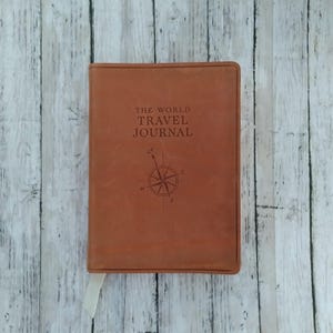 May include: A brown leather journal with the embossed text "THE WORLD TRAVEL JOURNAL" and a compass rose design. A white ribbon bookmark is visible. The journal is rectangular and appears to be closed.