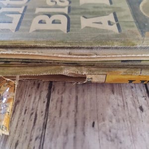 Pu&ograve; includere: Primo piano di un libro vintage intitolato "Little Black Sambo". La copertina &egrave; verde sbiadito con lettere bianche. Le pagine sono vecchie e usurate, con un bordo strappato. Il libro &egrave; appoggiato su una superficie di legno.