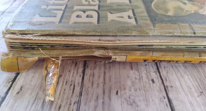 Pu&ograve; includere: Primo piano di un libro vintage intitolato "Little Black Sambo". La copertina &egrave; verde sbiadito con lettere bianche. Le pagine sono vecchie e usurate, con un bordo strappato. Il libro &egrave; appoggiato su una superficie di legno.