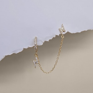 Può includere: Orecchino dorato con design a fulmine, ornato di piccole pietre scintillanti. L'orecchino presenta una catena che si collega a un secondo fulmine. Il nome del marchio "TINA KERR JEWELLERY" è stampato in basso.