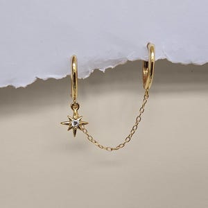 Peut inclure: Une boucle d'oreille double anneau dorée reliée par une fine chaîne. L'un des anneaux comporte une breloque en forme d'étoile avec une pierre centrale transparente. Le bijou porte l'inscription TIHA KERR JEWELLERY en bas de l'image.