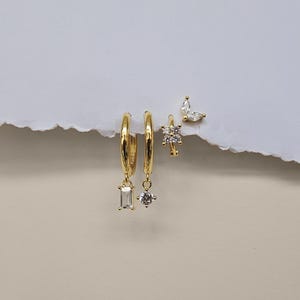 Peut inclure: Une collection de boucles d'oreilles dorées. Deux créoles avec des breloques pendantes, une rectangulaire et une en forme d'étoile, sont présentées avec deux petites boucles d'oreilles, une fleur et l'autre en forme de feuille. La marque "TINA KERR JEWELLERY" est en bas.