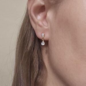 Può includere: Primo piano di un orecchino dorato con un piccolo diamante rotondo e un diamante pendente a forma di goccia. L'orecchino è indossato su un orecchio. Il marchio "TILA KERR JEWELLERY" è visibile in basso.