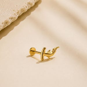 Pode incluir: Um brinco dourado em forma de tubarão-martelo com um fecho circular plano. A parte frontal em forma de tubarão tem um acabamento liso e polido, e a marca de metal S925 é visível na base circular. A peça é apresentada como uma unidade individual.