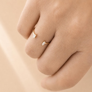 May include: An open gold ring featuring two small, clear baguette-cut stones at each end. The band is thin and polished, with the stones set horizontally. The text TINA KERR JEWELLERY is printed at the bottom of the image in a dark font.