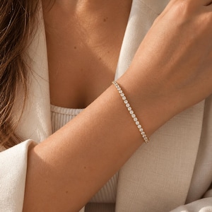 May include: A gold tennis bracelet featuring a continuous row of round, clear, brilliant-cut stones set in a polished metal band. The bracelet is worn on a wrist, showcasing the uniform size and sparkle of the stones against the gold setting.