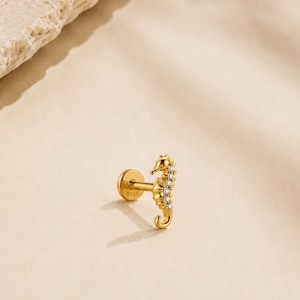 Peut inclure: Une boucle d'oreille clou en forme d'hippocampe doré avec un fermoir plat circulaire et une tige gravée S925. Le corps de l'hippocampe est orné d'une rangée verticale de petites pierres claires et rondes sur le bord avant, créant un design détaillé.