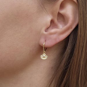 Op de afbeelding: Een gouden oorring met een kleine, getextureerde schelpbedel die onderaan hangt. Het midden van de schelpbedel is geaccentueerd met een minuscuul, rond, wit pareltje. De oorbel wordt in de oorlel gedragen en heeft een gepolijste afwerking.