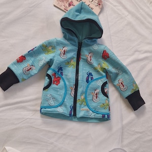 Könnte beinhalten: Eine hellblaue Kapuzenjacke mit schwarzem Reißverschluss und schwarzem Besatz. Die Jacke ist mit einem Muster aus bunten Drachen bedeckt.