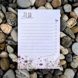 Könnte beinhalten: Ein weißer Notizblock mit den Worten "TO DO" in Schreibschrift oben. Der Notizblock hat linierte Seiten und ein Blumendesign am unteren Rand mit rosa und lila Blumen. Auf der rechten Seite des Notizblocks befindet sich ein kleiner Schmetterling.