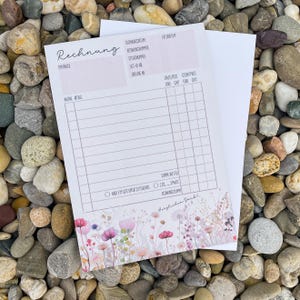 Könnte beinhalten: Eine weiße Rechnung mit dem Wort "Rechnung" oben links. Die Rechnung hat ein Blumendesign am unteren Rand mit rosa und lila Blumen. Die Rechnung liegt auf einem Bett aus kleinen, glatten Steinen.