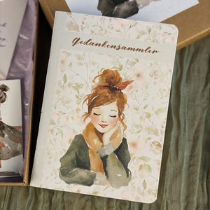 Könnte beinhalten: Ein Notizbuch mit einer Aquarellillustration einer Frau mit rotbraunem Haar, die einen Schal und einen Mantel trägt. Das Notizbuch hat einen floralen Hintergrund und den deutschen Text "Gedankensammler". Das Notizbuch befindet sich in einer Schachtel mit passender Illustration.