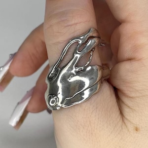 May include: A silver-colored ring with an abstract, organic design. The ring features flowing, melted-like shapes and a smooth, polished finish. The ring is worn on a finger.