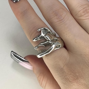 May include: A silver-colored ring with an abstract, melted design. The ring has a unique, flowing shape with multiple protrusions and a smooth, reflective surface. The ring is worn on a finger, showcasing its sculptural form and modern aesthetic.