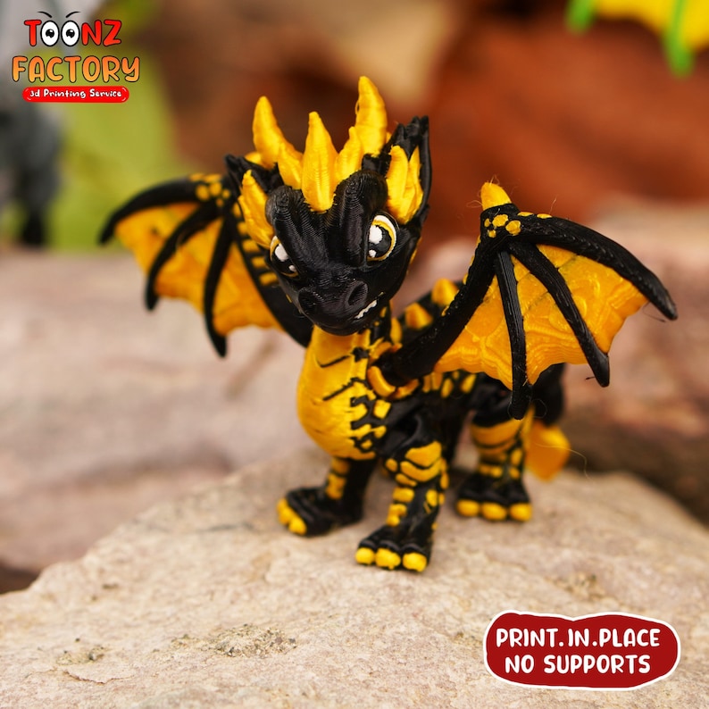 K&ouml;nnte beinhalten: Eine 3D-gedruckte Drachenfigur in Schwarz und Gelb mit ausgebreiteten Fl&uuml;geln. Der Drache hat einen stacheligen gelben Kamm und gro&szlig;e, runde Augen. Der Text "PRINT.IN.PLACE NO SUPPORTS" ist auf der Basis aufgedruckt.
